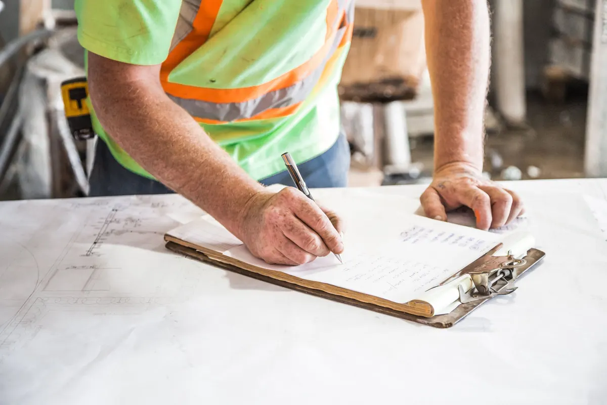 A worker in a high-visibility shirt writes on a clipboard resting on a set of building plans.