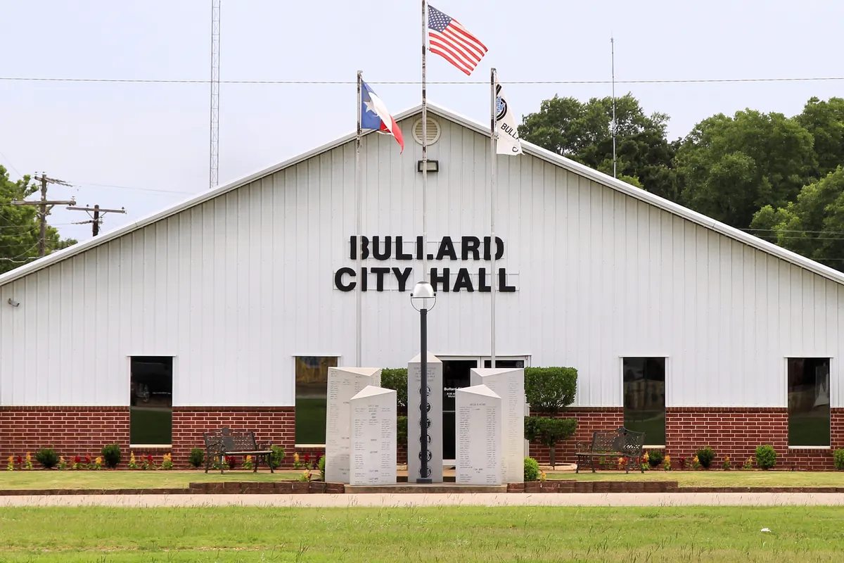 Bullard City Hall, a white metal building with a brick base, with flags and a stone monument out front.