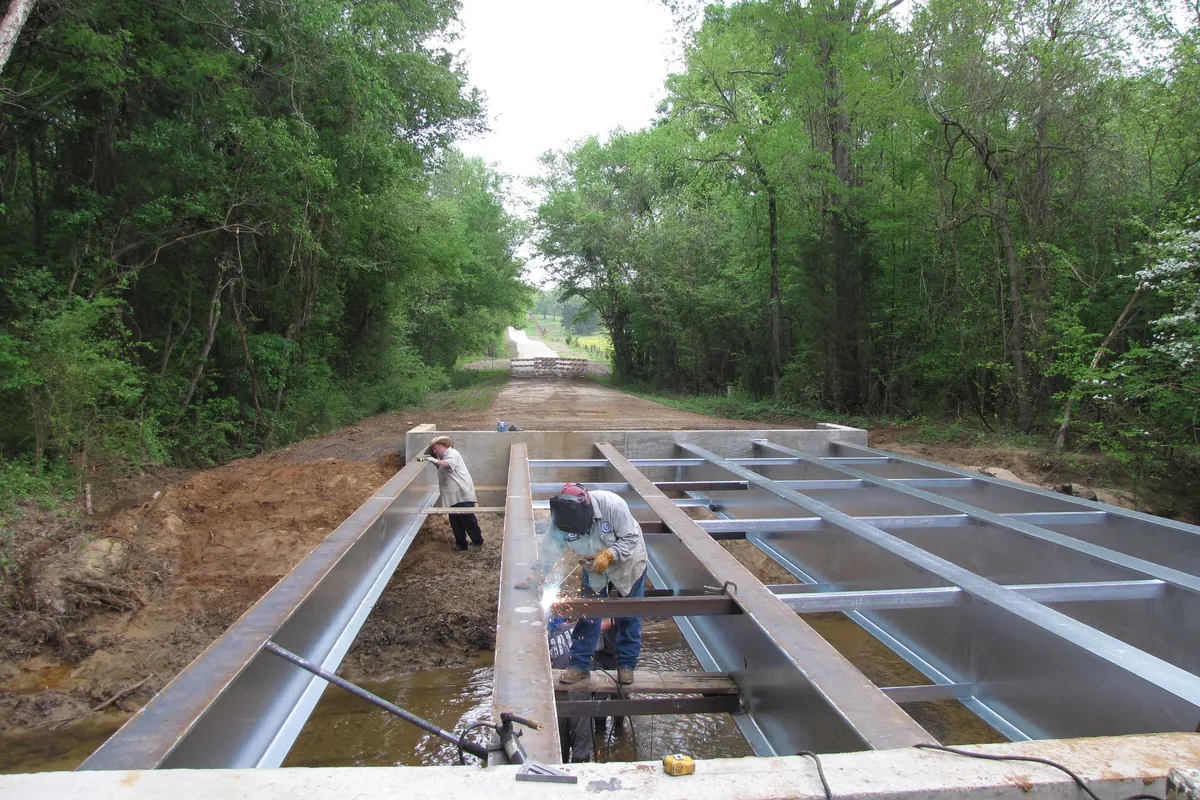 Workers assembling the steel frame of a small bridge on a dirt county road through the woods.
