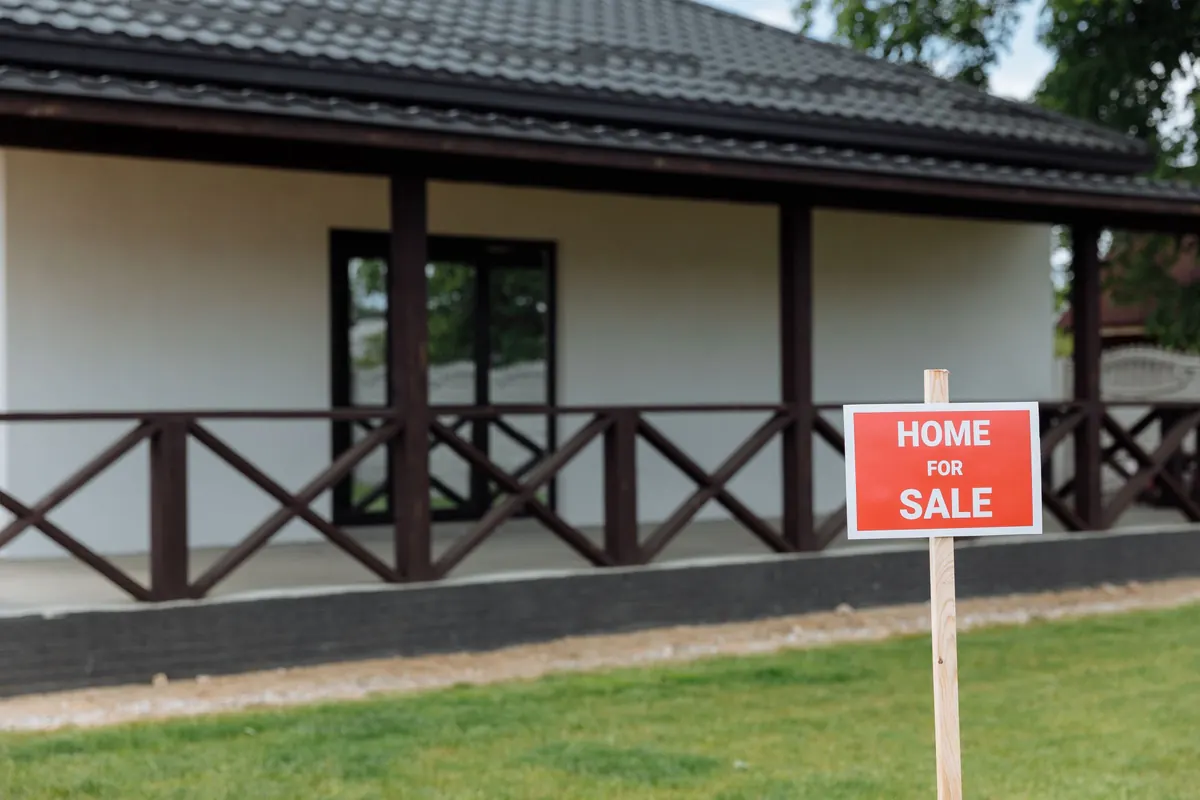 A red Home for Sale sign on a post in front of a single-story house with a covered porch.