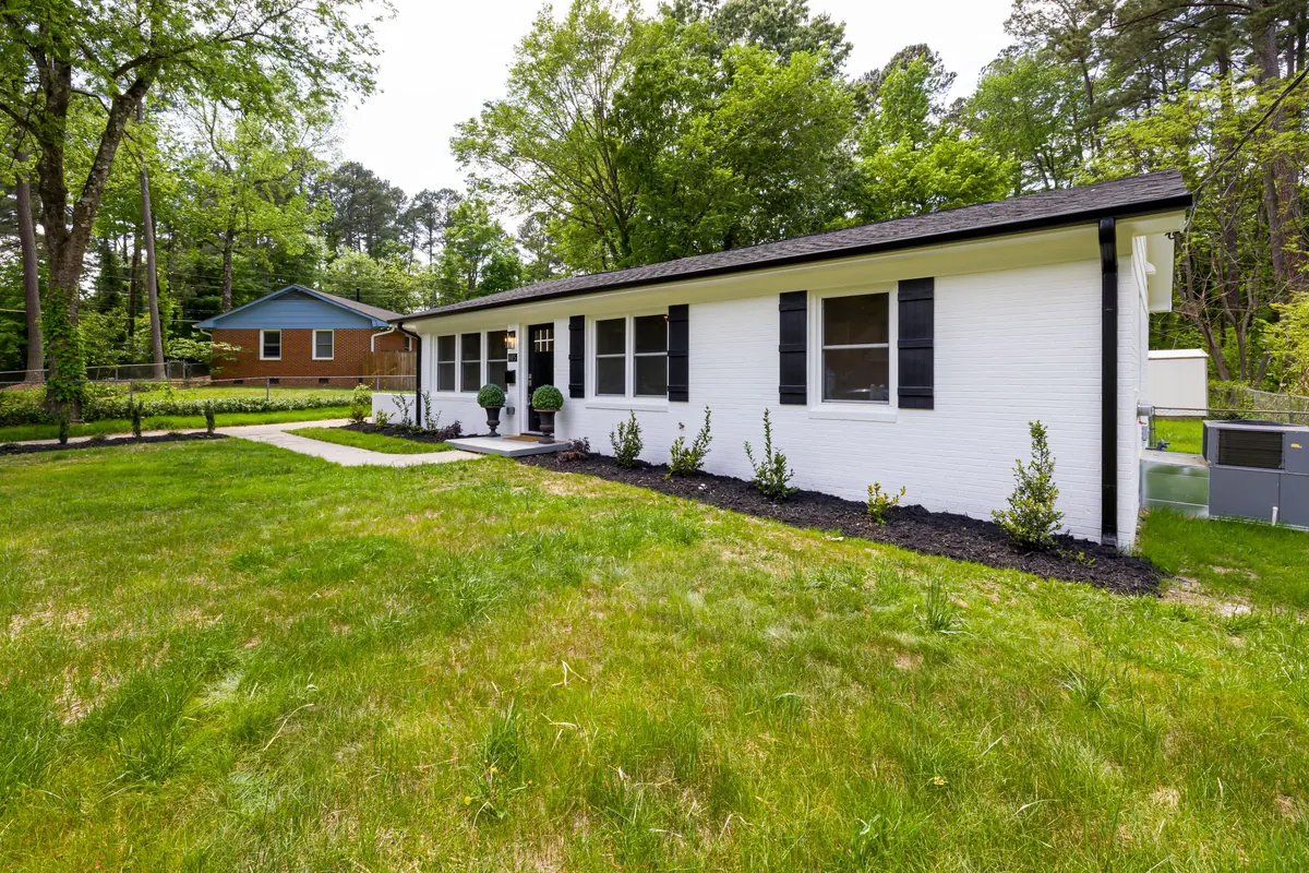 A long single-story ranch house of white-painted brick with black shutters, set on a wide green lawn with tall trees behind it.