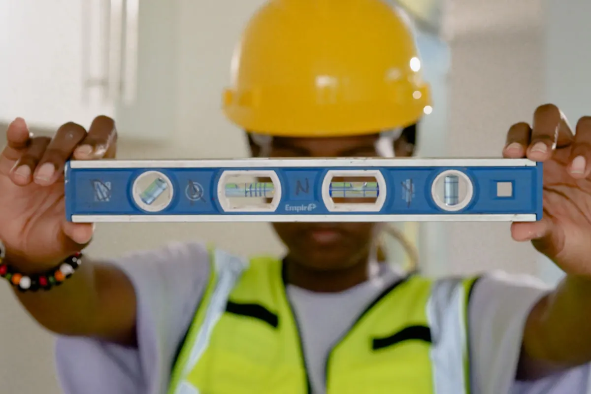 A worker in a yellow hard hat and safety vest holds a long spirit level up at eye level.