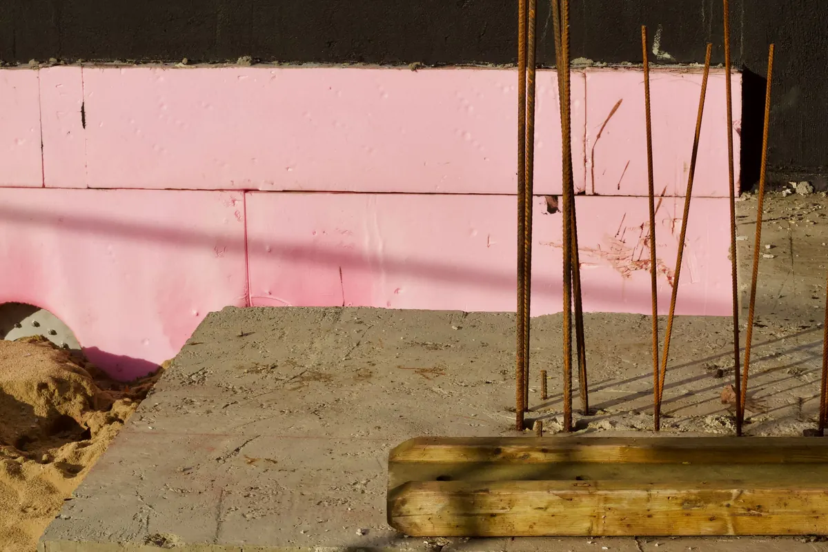Steel rebar rods rising from the edge of a concrete slab in front of pink foam insulation board, with a wooden form board beside them.