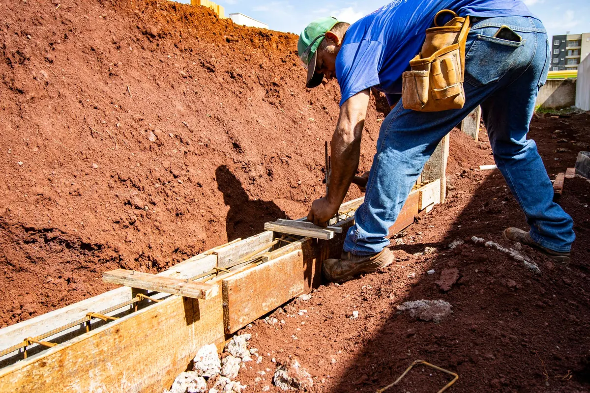 A worker with a tool belt sets wooden forms in a trench cut into red clay soil.