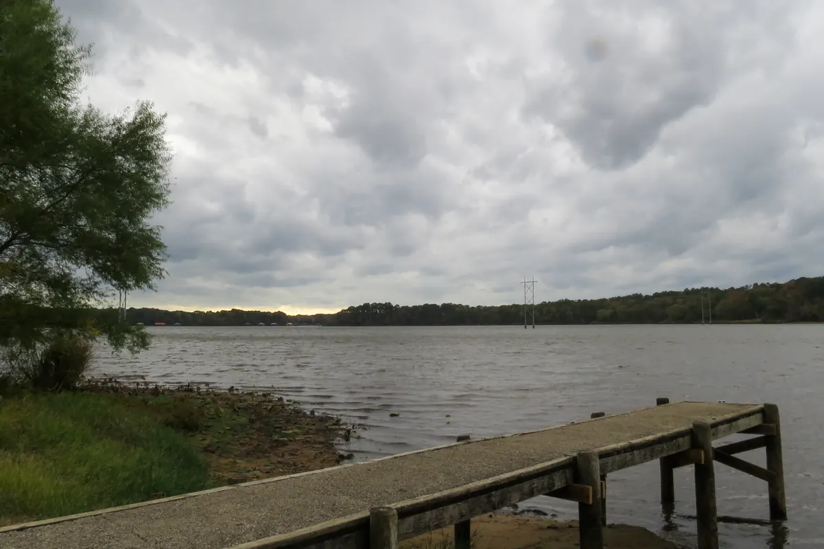 A concrete fishing pier reaching into Lake Tyler East under a cloudy sky, with a wooded shoreline across the water.